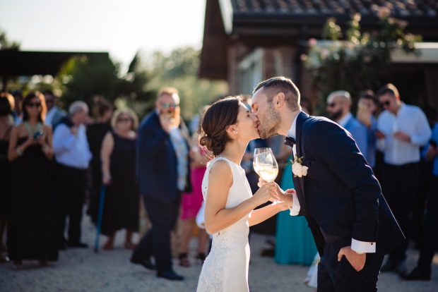 Gli sposi si scambiano un bacio romantico brindando con Franciacorta, mentre gli invitati festeggiano sullo sfondo.