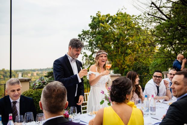 Gli sposi brindano sorridenti al tavolo sulla terrazza di Villa Calchi, celebrando il loro matrimonio. Fotografo matrimonio Varese Villa Calchi.
